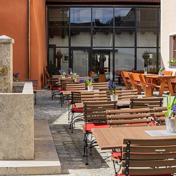 Der Biergarten der Gastwirtschaft Schattenhofer  ist umgeben von den Wänden und dem Neubau im Hintergrund aus Glas. Links ist ein Steinbrunnen zu sehen.