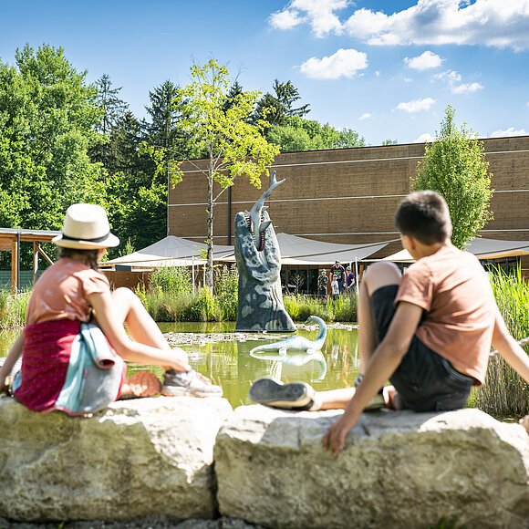 Zwei Kinder sitzen am Wasser im Außengelände des Dinosauriermuseum und beobachten die grüne Landschaft und das Wasser.