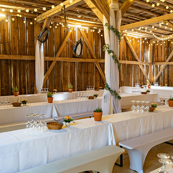 Zwei lange Tafeln mit weißer Tischdecke stehen unter einem Holzdach, auch die Seiten sind mit Holz vor Wind und Wetter geschützt.