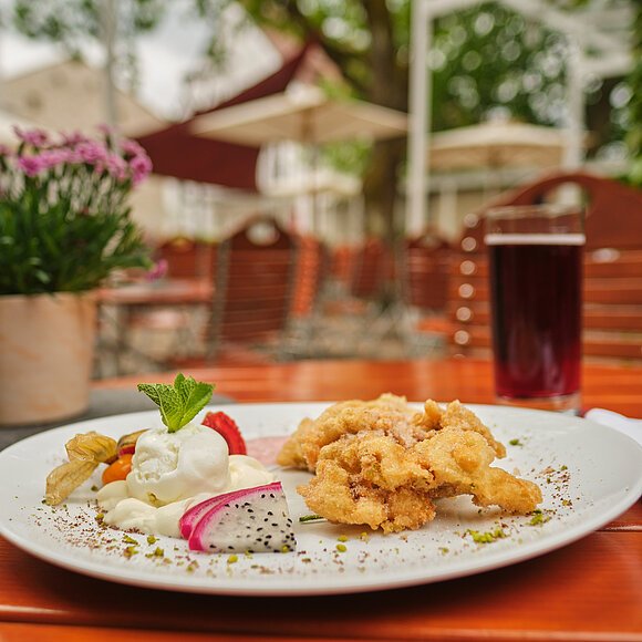 Auf einem Holztisch auf der Terrasse ist eine köstlich angerichtete Portion Obatzda serviert. Im Hintergrund stehen links zwei kleine Blumentöpfe und rechts ein Getränk.