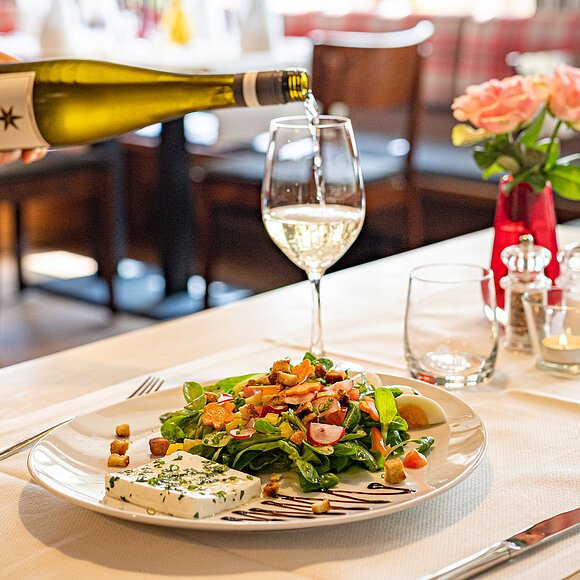 Eine Hand gießt gerade Wein aus einer Flasche in ein Weinglas ein. Im Vordergrund steht ein Teller mit Salat und Käse.