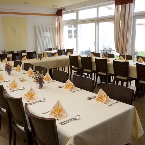 Ein großer Raum mit gedeckten Tischen, Stühlen und gefalteten Servietten, Fenster im Hintergrund.