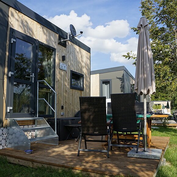 Moderne Holzhütte mit Terrasse, Tisch, Stühlen und Sonnenschirm im Freien, umgeben von Bäumen.