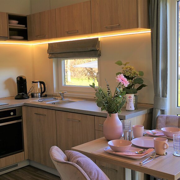 Kleine Küche mit Holzschränken, Elektrogeräten, Spüle und Esstisch. Fenster mit Blick ins Freie.