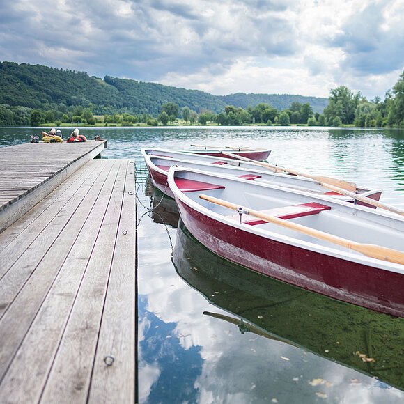 Zwei Ruderboote sind an einem Holzsteg festgemacht, umgeben von einem See und bewaldeten Hügeln.