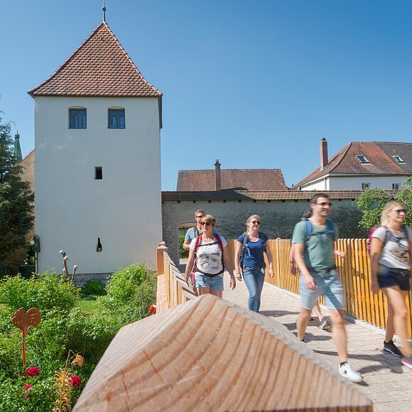 Fünf Personen mit Rucksäcken gehen auf einem gepflasterten Weg neben einem weißen Turm mit rotem Dach bei klarem Himmel.