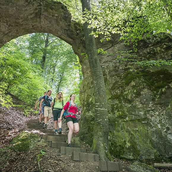 Vier Wanderer gehen einen Waldweg mit Steintreppe unter einem natürlichen Felsbogen hinab.