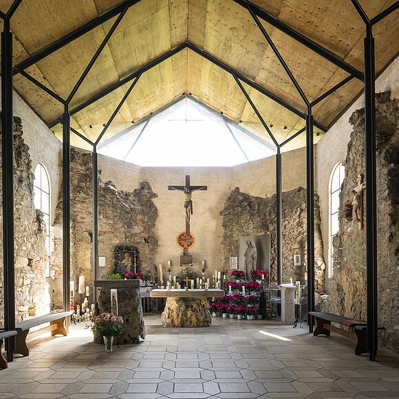 Innenraum einer Kapelle mit Altar, Kreuz, Kerzen und Blumen, umgeben von alten Steinmauern und Holzdecke
