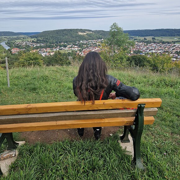 Person mit langen dunklen Haaren sitzt auf einer Bank und blickt auf eine Stadt im Tal unter bewölktem Himmel.