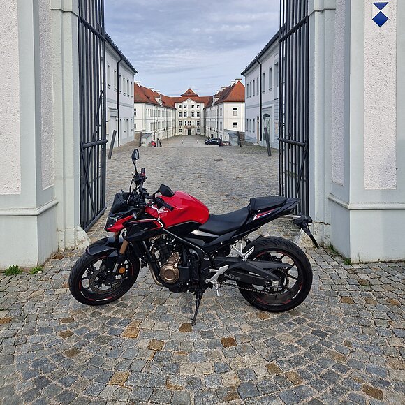 Motorrad vor dem Schloss Hirschberg Rotes Motorrad steht auf gepflastertem Hof zwischen geöffneten schwarzen Toren vor weißen Gebäuden mit roten Dächern.