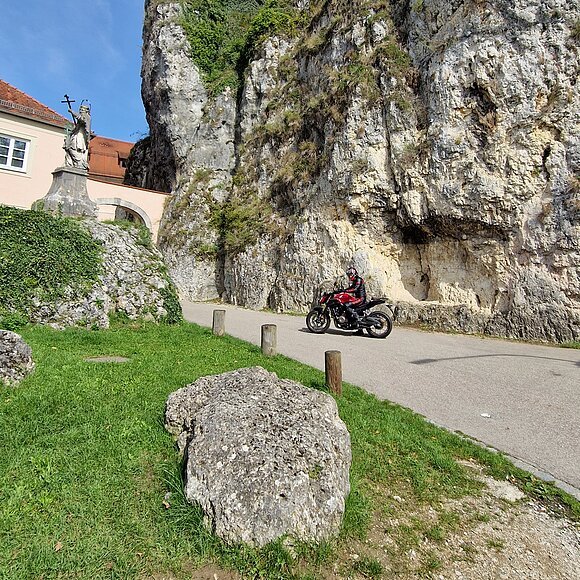 Motorradfahren in Weltenburg Motorradfahrer auf Straße vor steiler Felswand, links Statue und Gebäude mit rotem Dach.