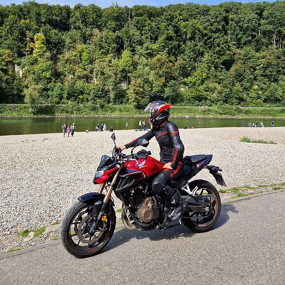 Motorradfahren in Weltenburg Person in schwarzer Motorradkleidung und Helm auf rotem Motorrad auf Weg am Flussufer mit Wald im Hintergrund