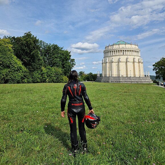 Motorrradfahrerin an der Befreiungshalle Kelheim Person in schwarzer Motorradkleidung mit Helm in der Hand steht auf Wiese vor rundem Gebäude mit Säulen und Statuen.