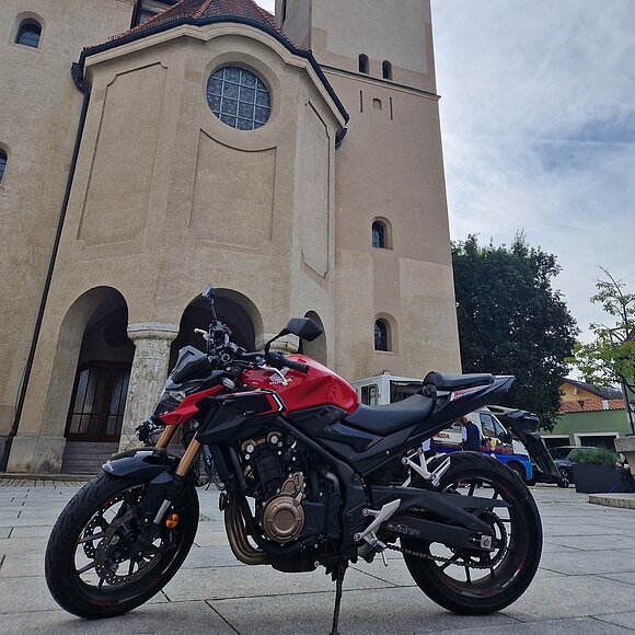 Motorrad vor der Kirche Hl. Walburga in Beilngries Rotes Motorrad steht vor einer Kirche mit zwei Türmen und einem Uhrenturm bei bewölktem Himmel.