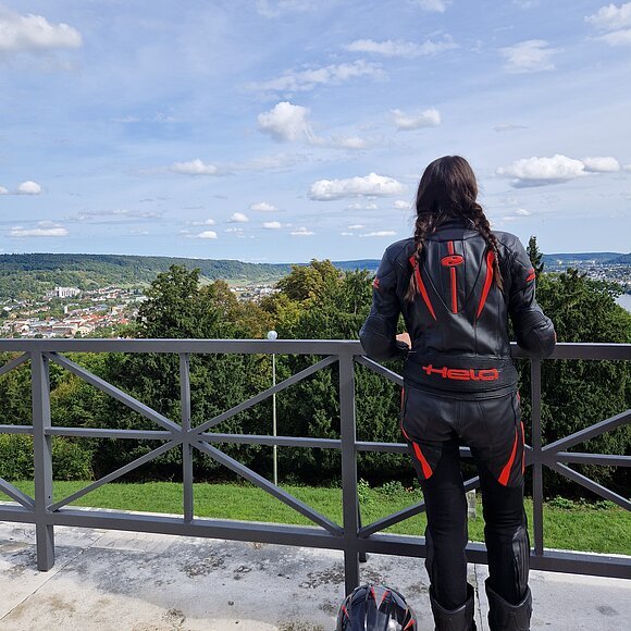 Person in schwarzer Motorradkleidung mit roten Akzenten blickt von Balkon auf grüne Landschaft und Stadt, Helm liegt am Boden.