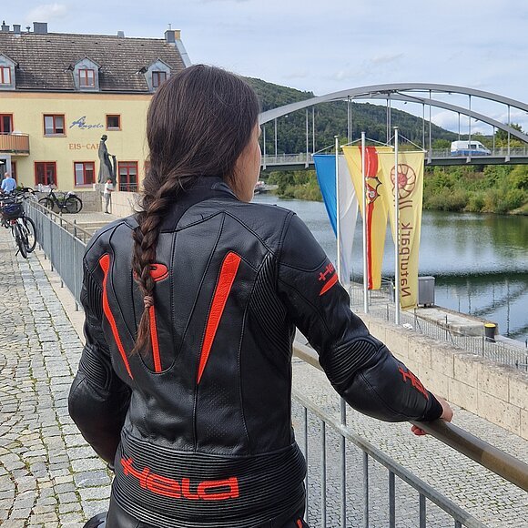 Person mit geflochtenem Haar und schwarzer Motorradjacke mit roten Akzenten lehnt an Geländer am Flussufer mit Brücke und Gebäuden im Hintergrund.