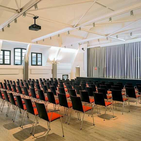 Leerer Konferenzraum mit Reihen schwarzer Stühle mit roten Sitzflächen vor einer Bühne mit grauem Vorhang.