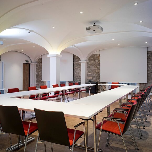 Konferenzraum mit U-förmigen weißen Tischen, roten Stühlen und gewölbter Decke mit Steinwand im Hintergrund.