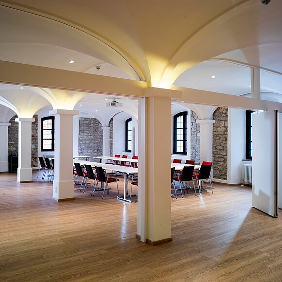 Großer heller Raum mit Holzboden, weißen Säulen, Tischen und Stühlen, teils durch Trennwände geteilt.