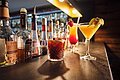 Bar mit verschiedenen Spirituosenflaschen und drei Cocktails auf der Theke, darunter ein brauner Drink mit Zimtstange.
