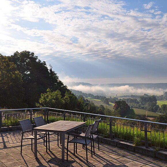 Terrasse mit Tisch und Stühlen, Blick auf neblige Landschaft mit Bäumen und Feldern bei bewölktem Himmel.