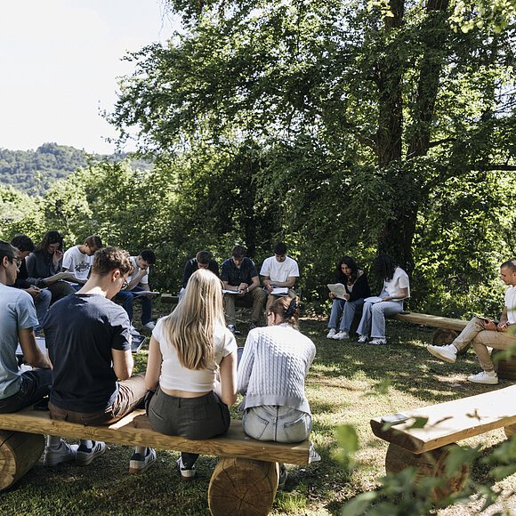 Gruppe von Menschen sitzt im Kreis auf Holzbänken im Freien und liest oder schreibt.