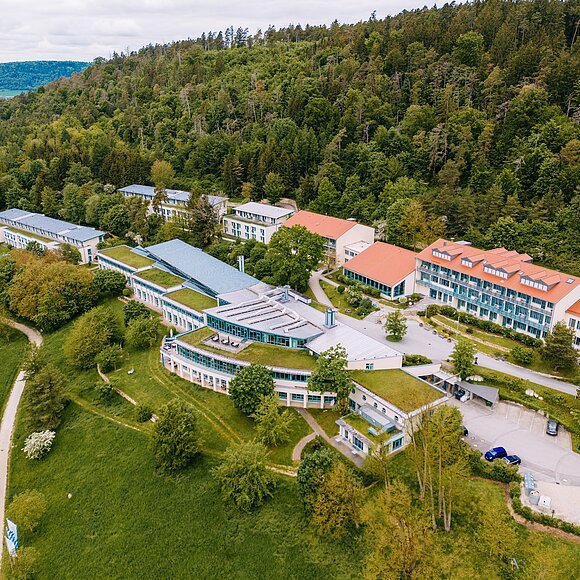 Luftaufnahme eines Gebäudekomplexes mit grünen Dächern am Waldrand und umliegenden Wiesen und Wegen.