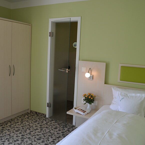 Hotelzimmer mit Einzelbett, grün gestrichener Wand, Schrank, Glastür und Nachttisch mit Lampe und Blumen.