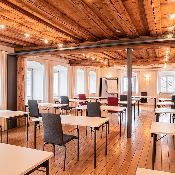 Seminarraum mit Holzboden, Holzbalkendecke, weißen Tischen und roten sowie grauen Stühlen, Fenster mit weißen Vorhängen.