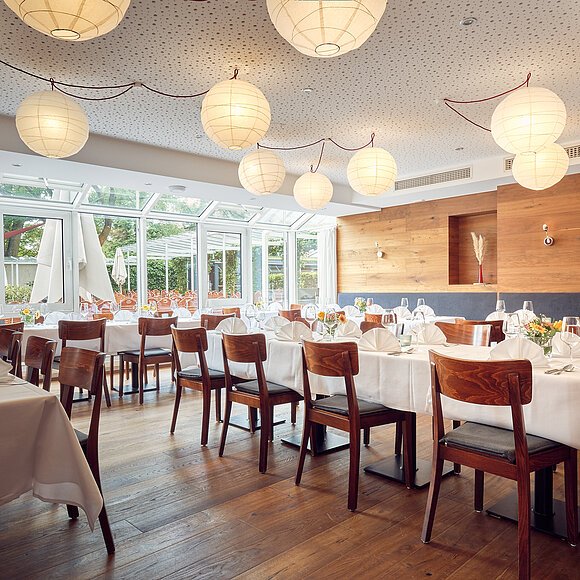 Restaurant mit gedeckten Tischen, Holzstühlen, großen Fenstern und runden Papierlampen an der Decke.