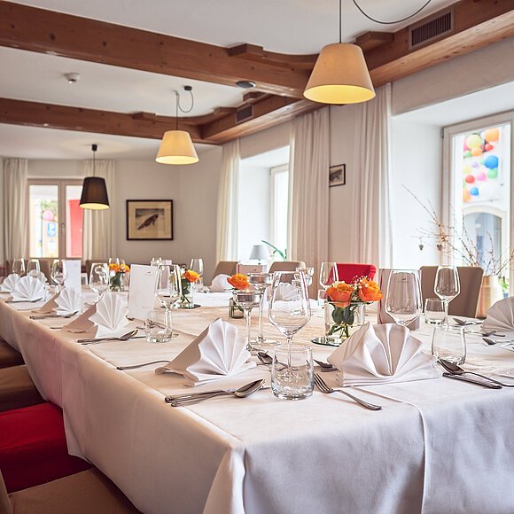 Großer Esstisch mit weißer Tischdecke, gefalteten Servietten, Gläsern und Blumen in einem Raum mit Holzdecke.
