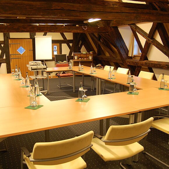 Konferenzraum mit U-förmigen Tischen, Stühlen, Wasserflaschen und Flipcharts unter Holzbalken.