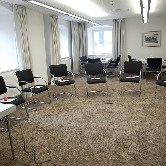 Konferenzraum mit Stühlen im Halbkreis, auf jedem Stuhl ein Apfel und ein Zettel, Laptop auf Tisch links.