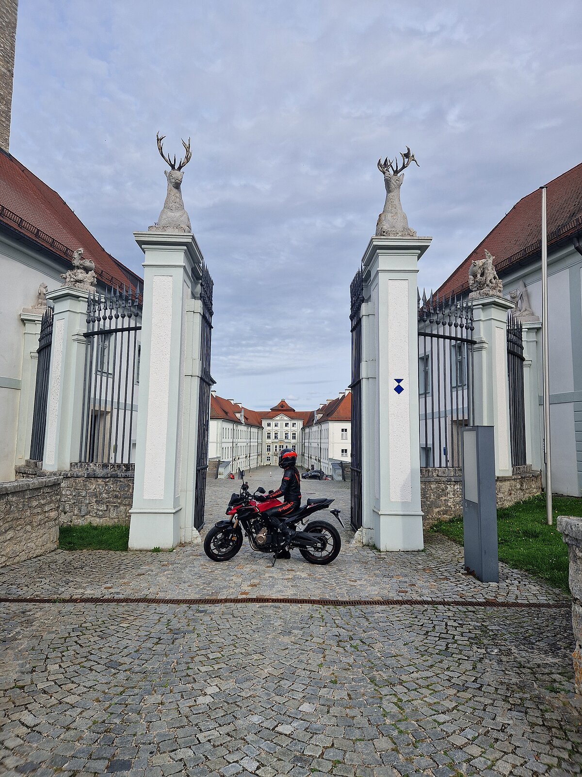 Motorradfahrerin vor dem Schloss Hirschberg Motorradfahrer in schwarzer und roter Schutzkleidung vor Tor mit Hirschstatuen und historischem Gebäude im Hintergrund.