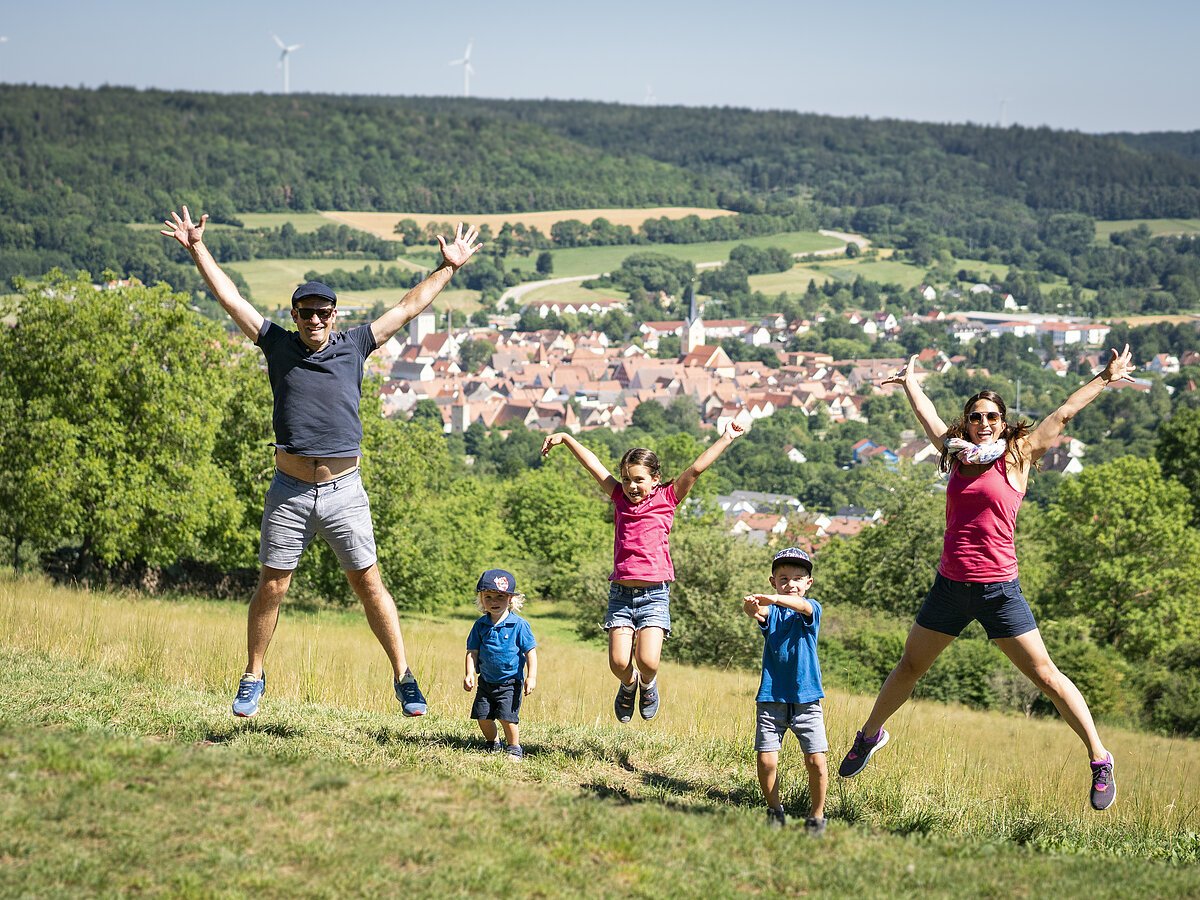 Eine vierköpfige Familie springt mit ausgestreckten Armen und Beinen oberhalb von Berching in die Höhe. Sie landen sanft auf einer grünen Wiese.