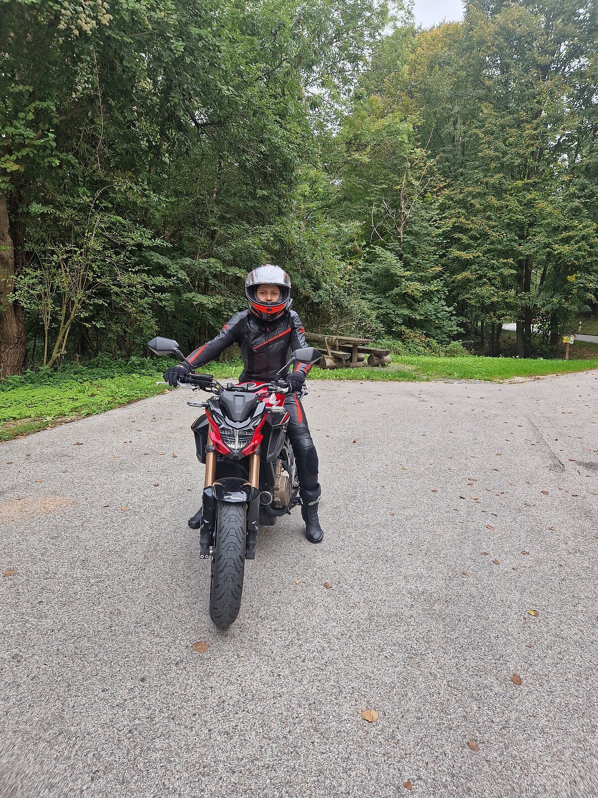 Motorradfahrerin Person in schwarzer Motorradkleidung und Helm sitzt auf rotem Motorrad auf asphaltierter Fläche vor Wald und Picknickbank.