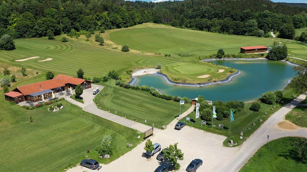 Eine Luftaufnahme von oben auf ein Haus mit See und viel grüner Wiese zum Golf spielen.