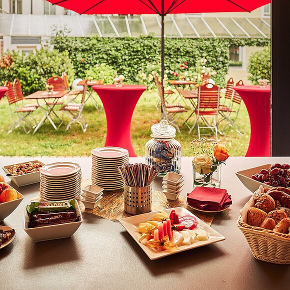 Kaffeepausenstation mit Blick in den Garten im Hotel Fuchsbräu Buffet mit Obst, Gebäck, Nüssen und Tellern auf Tisch vor Garten mit roten Stehtischen und Stühlen